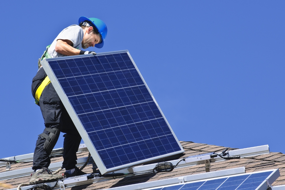 A technician wearing a blue hard hat and safety harness installs a solar panel on a residential shingle roof under a clear blue sky.
